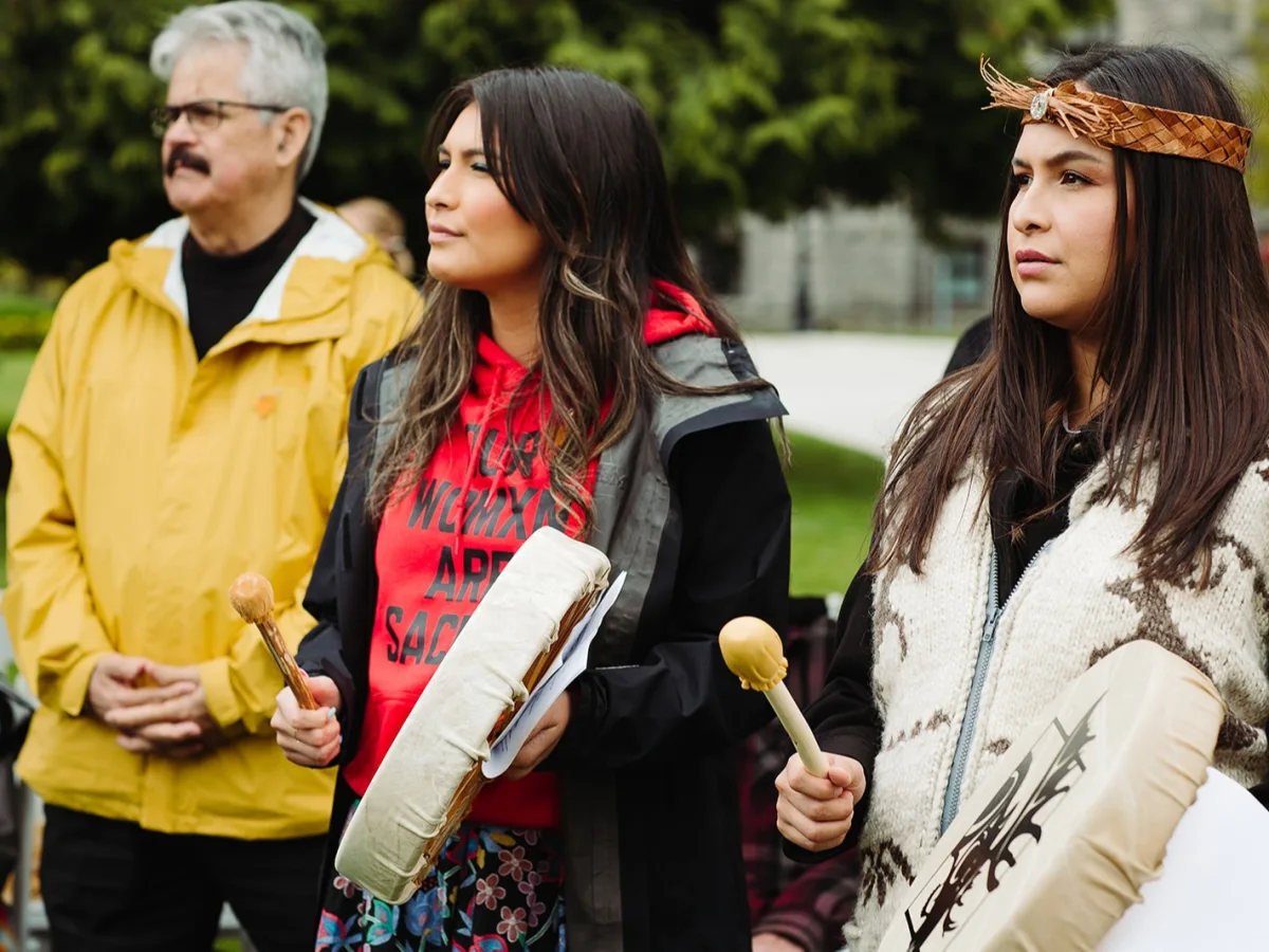 A man and two women standing outside during Moose Hide Campaign Day. Both women are holding traditional hand drums. 
