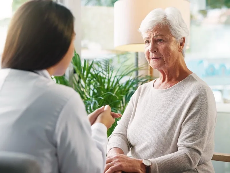 A healthcare professional in a white coat holds an older woman's hand during a conversation in a home setting.