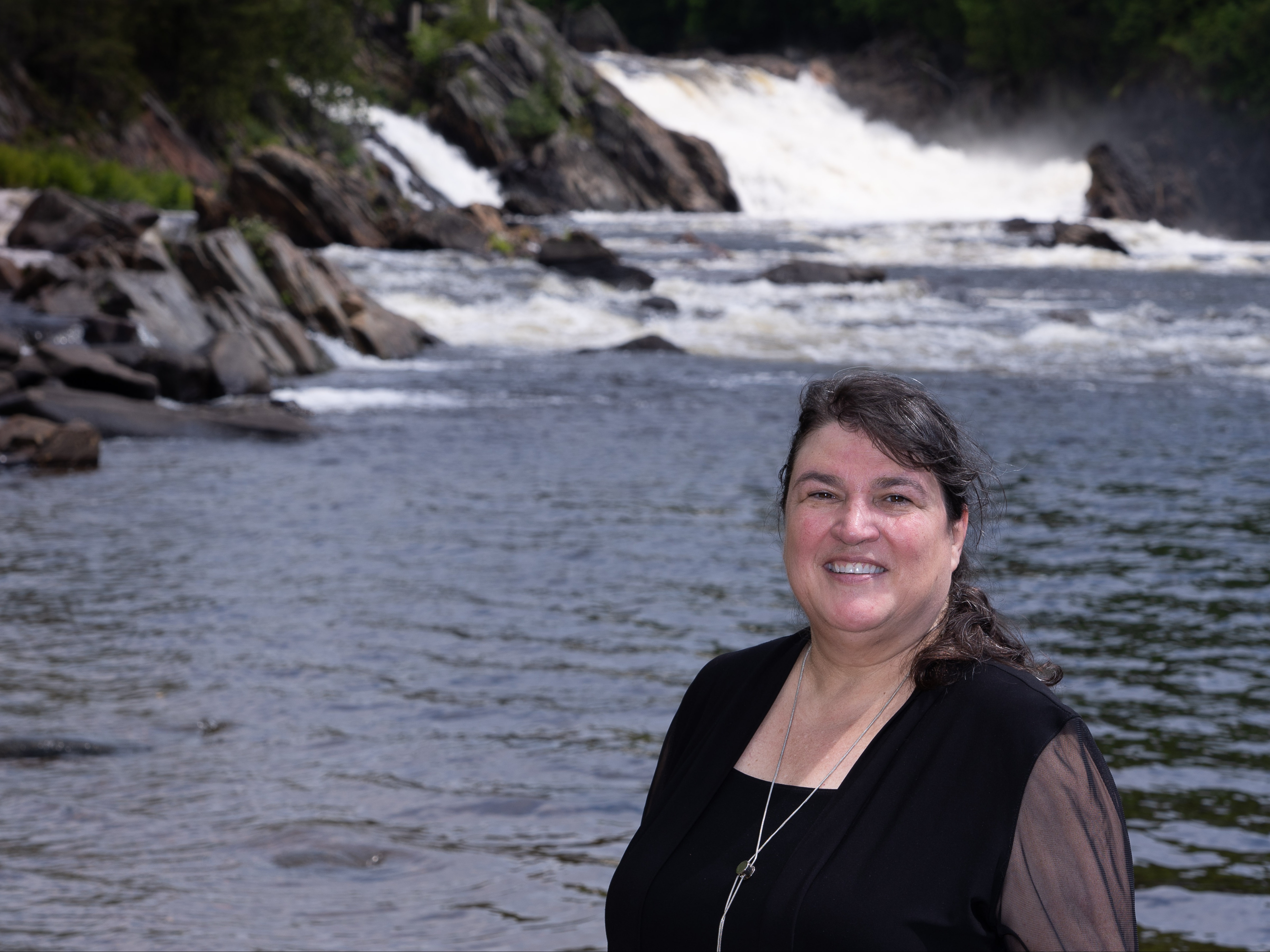 Woman smiling in front of a river with a small waterfall in the background.