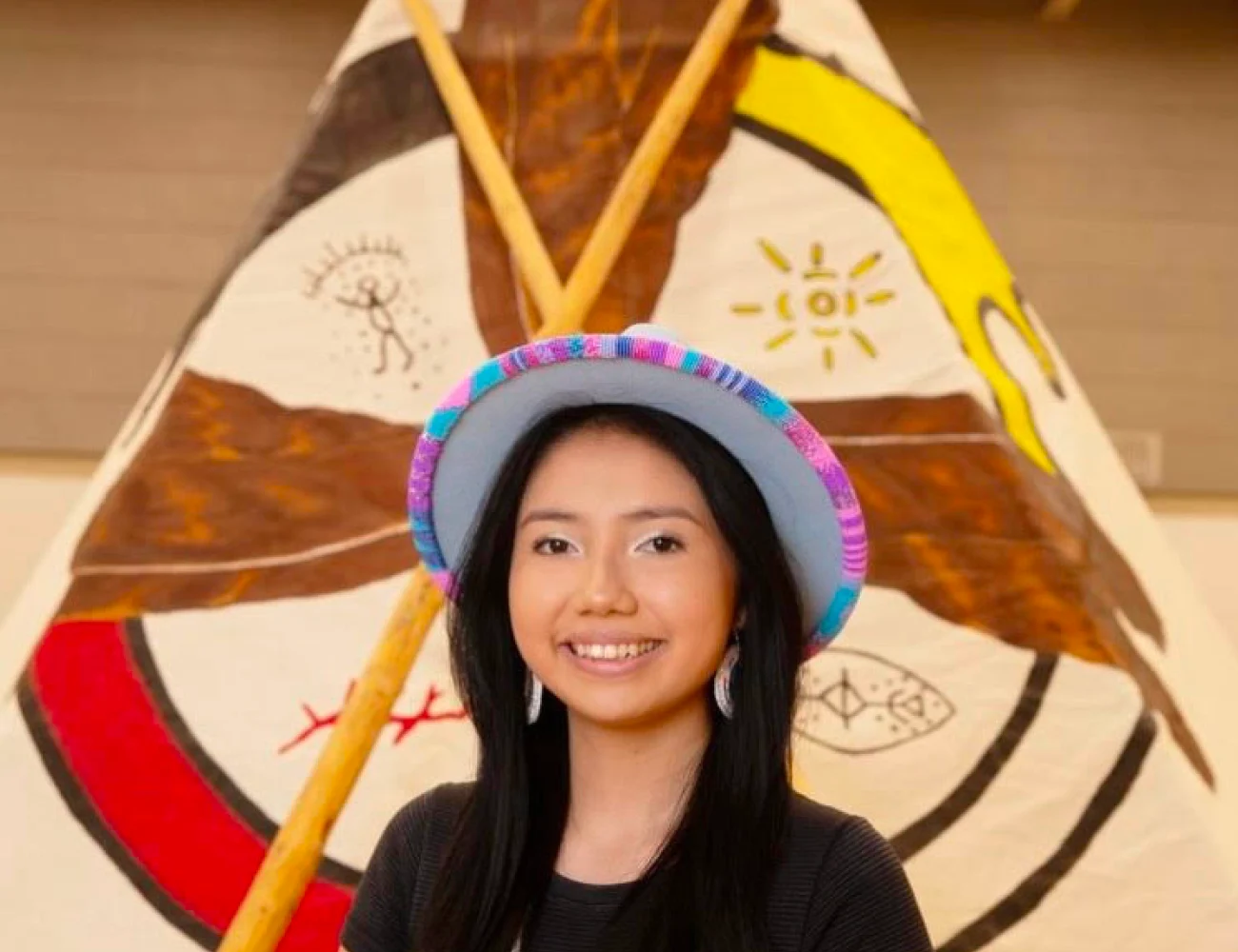 TELUS Student Bursary Recipient, Tiya, standing in front of a medicine wheel painting on a tipi at her Indigenous graduation ceremony.