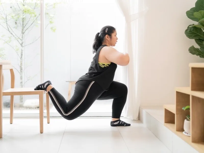 Femme effectuant une fente bulgare en utilisant une chaise en bois pour le soutien dans un espace de fitness minimaliste et lumineux avec des plantes