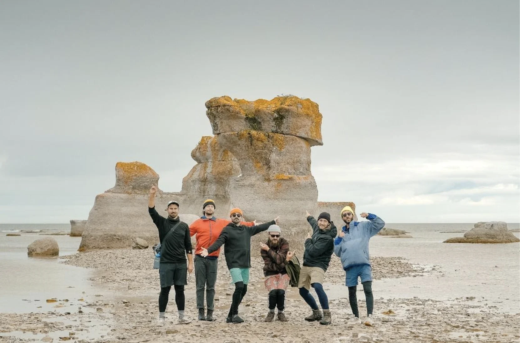 Members of Qualité Motel posing in front of a large rock on a beach