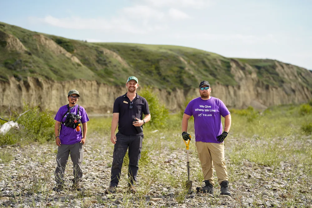 Three field workers standing in a rocky riverbed near grassy hills.