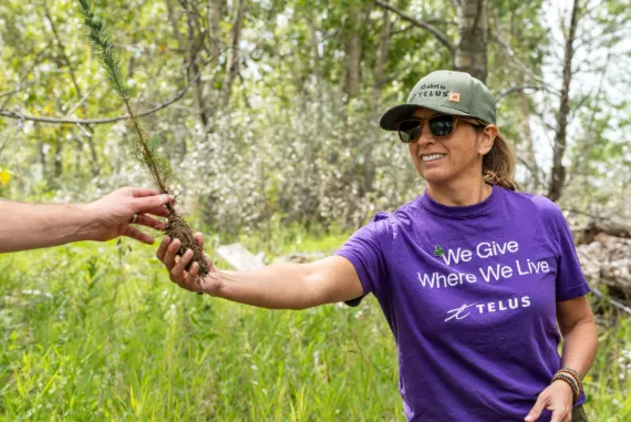 Une femme portant un chandail TELUS mauve remet un petit plant d’arbre à quelqu’un à l’extérieur.