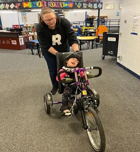 Educator pushing girl on her modified bike in a a school hallway.