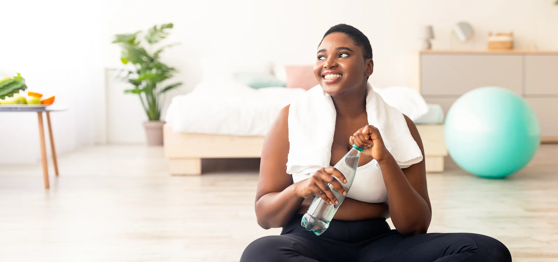 Woman sitting on exercise mat at home holding a water bottle, with dumbbells and fitness equipment nearby.