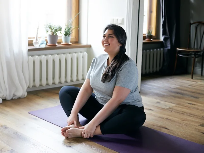 Smiling woman in gray shirt sitting cross-legged on a purple yoga mat in a bright bedroom, surrounded by plants and natural light