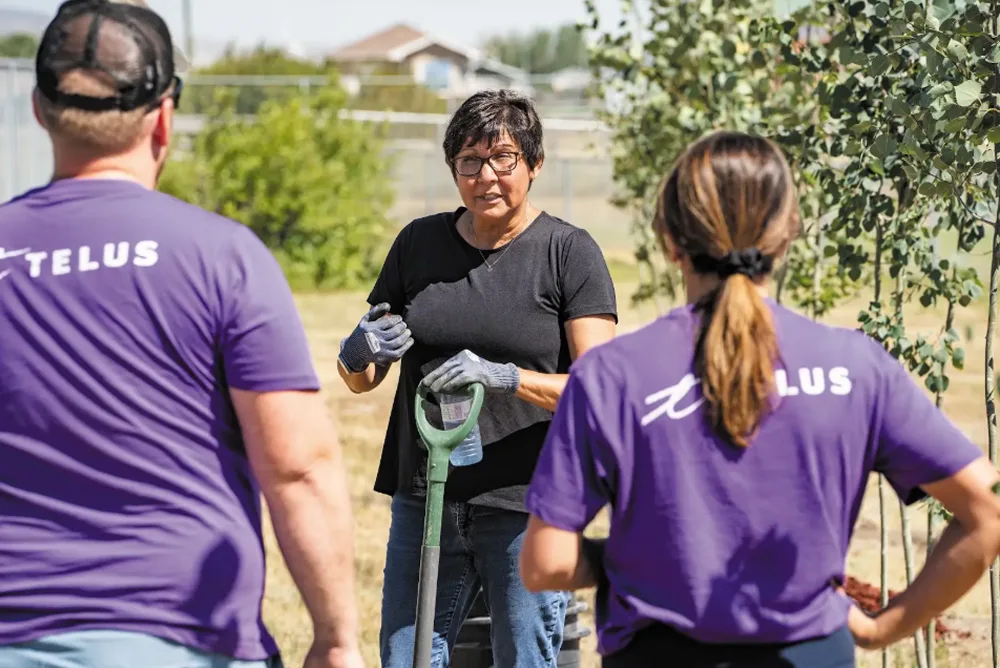 Woman holding a shovel speaking to two TELUS volunteers wearing purple shirts outdoors.