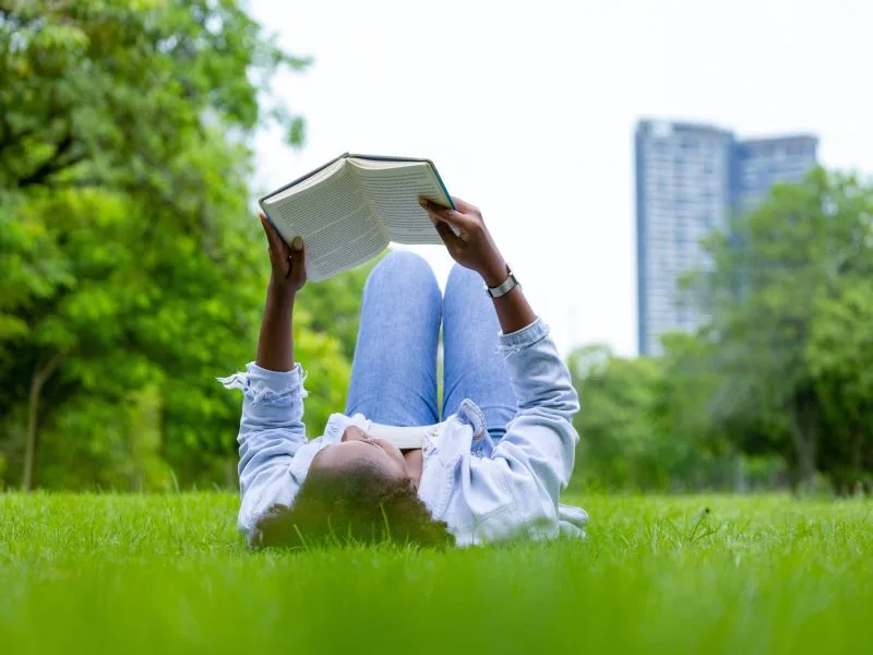 A woman lying down on a field in a public park while holding up and reading a book.