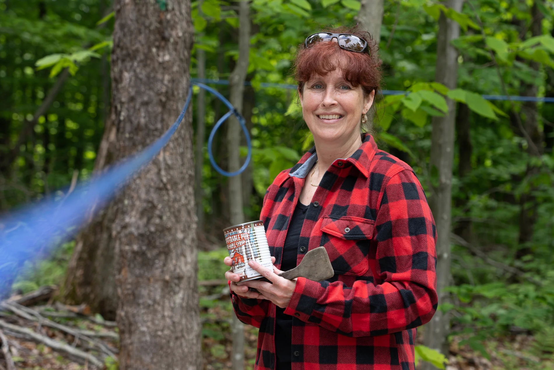 Woman smiling and holding a can of maple syrup in front of maple trees. 