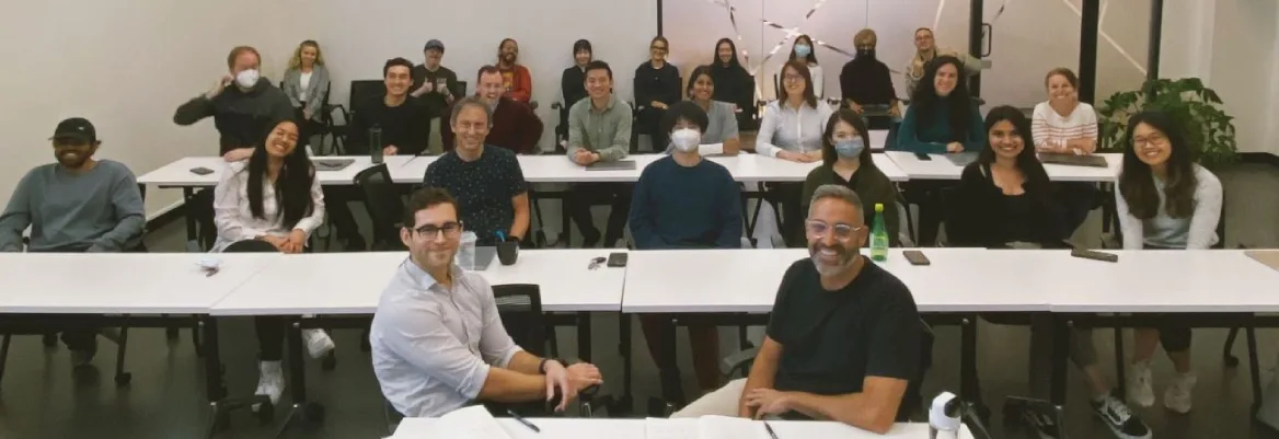 Several people, some wearing face masks, sit behind long white desks, looking toward the camera and smiling