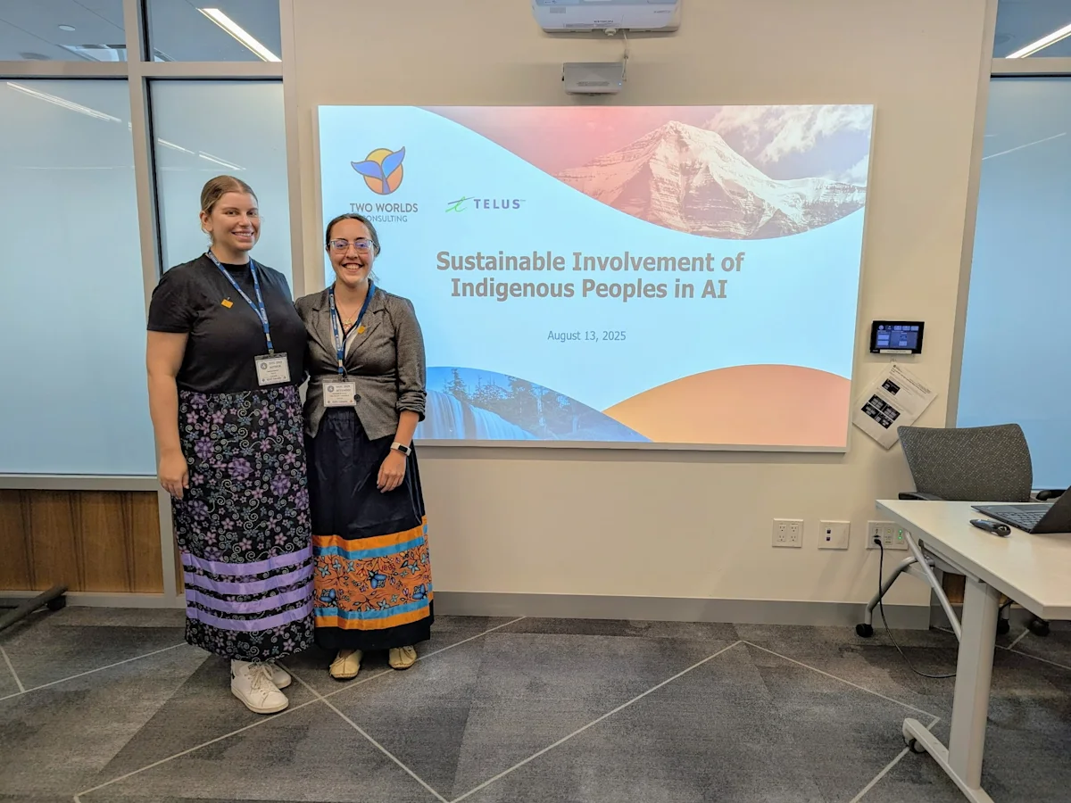 Kendal Burtch and Melanie Demers stand smiling in front of a projector showing a slide on Indigenous Peoples in AI. 

