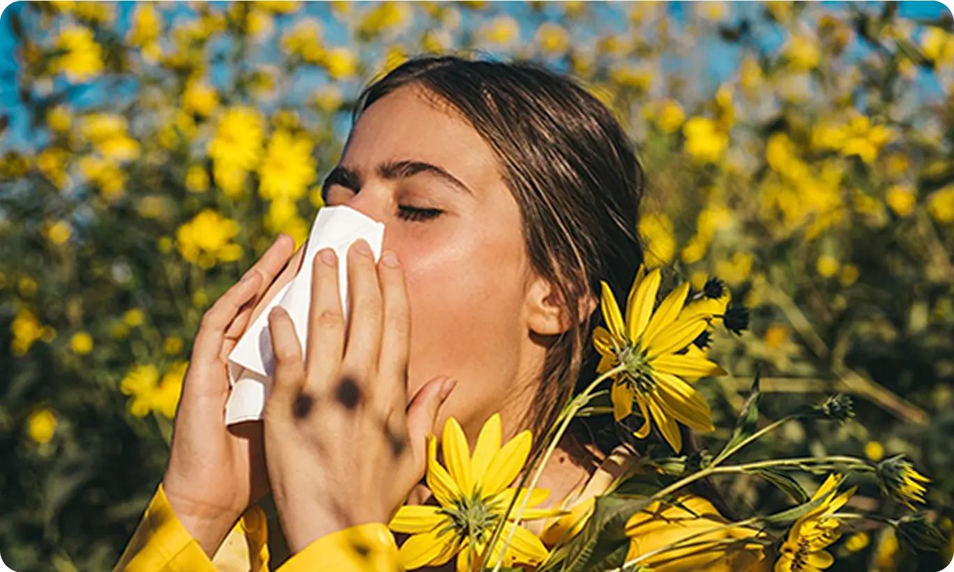 A woman holding a tissue in a field of yellow flowers, showing that allergies are in everyday life.