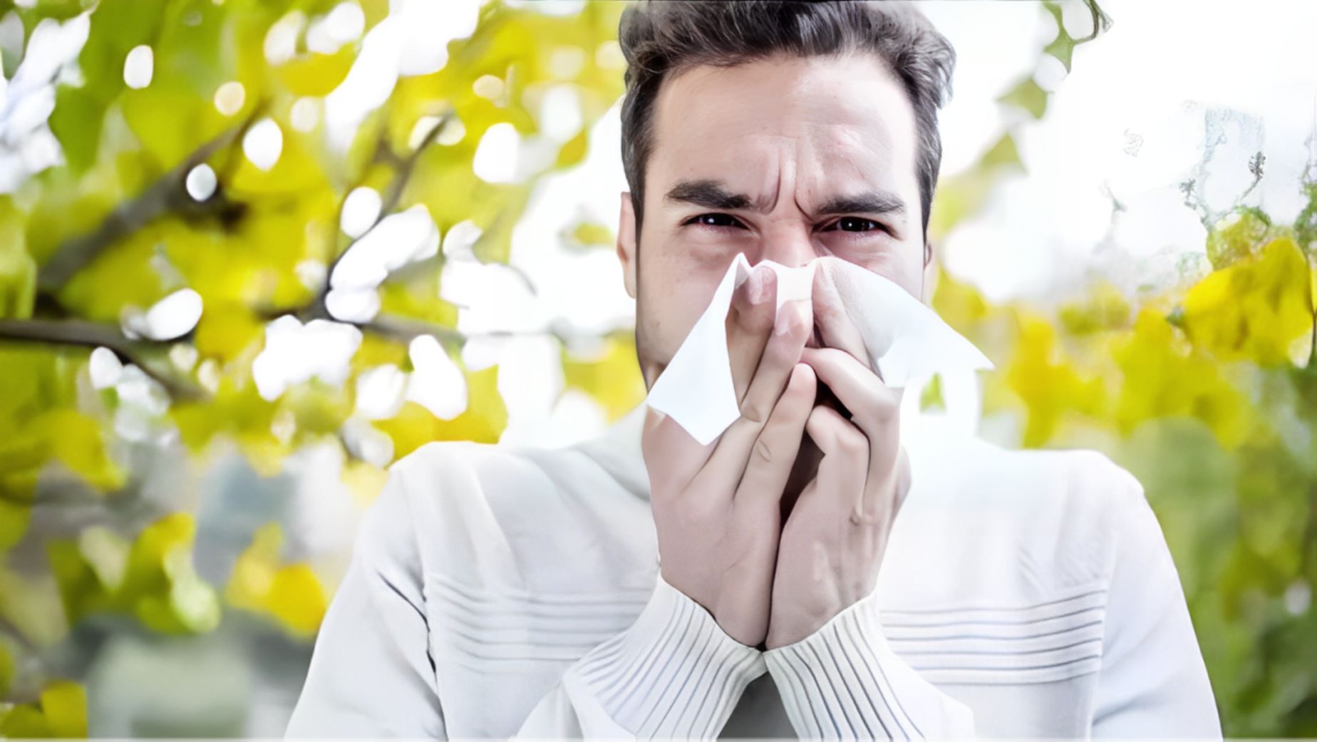 A man holding a tissue near his face surrounded by foliage showing discomfort of seasonal allergies.