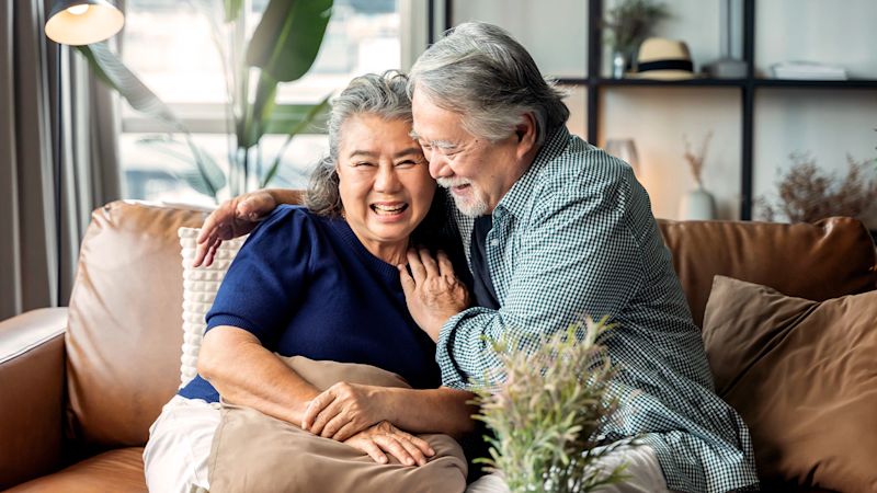 Elderly couple sitting on a brown leather sofa, smiling with the man in a blue checkered shirt and the woman in a navy shirt, surrounded by a lamp and houseplant.