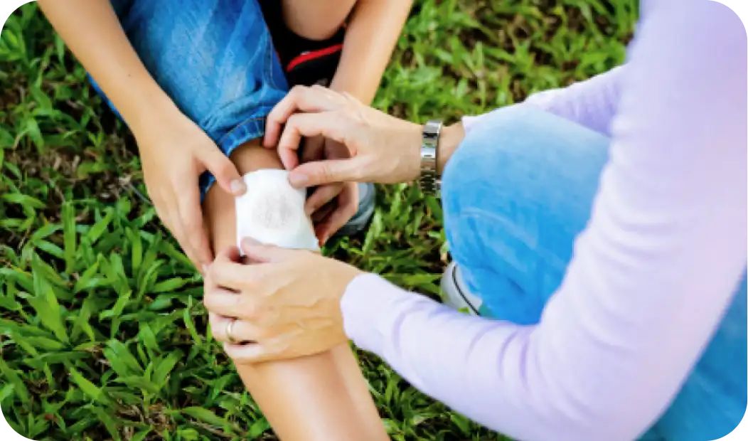 A person is gently placing a bandage on a child's scraped knee while they sit on grass, providing first aid care.