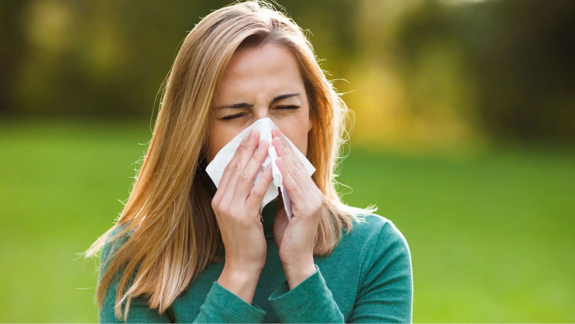 A woman blowing her nose into a tissue in an outdoor setting, showing discomfort of allergy symptoms