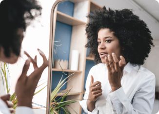 A woman with curly hair looking in a mirror and applying lip balm to treat chapped lips.