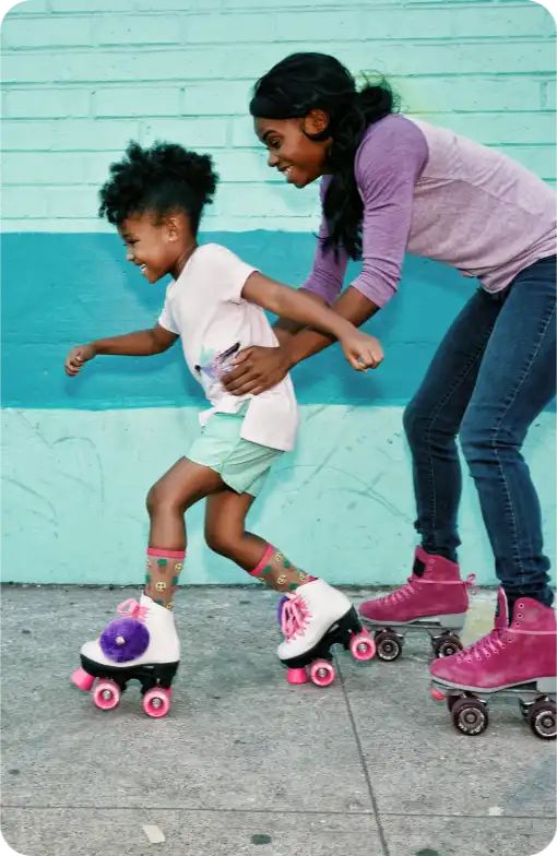 A woman and a child roller skating together on a sidewalk, both smiling and having fun.