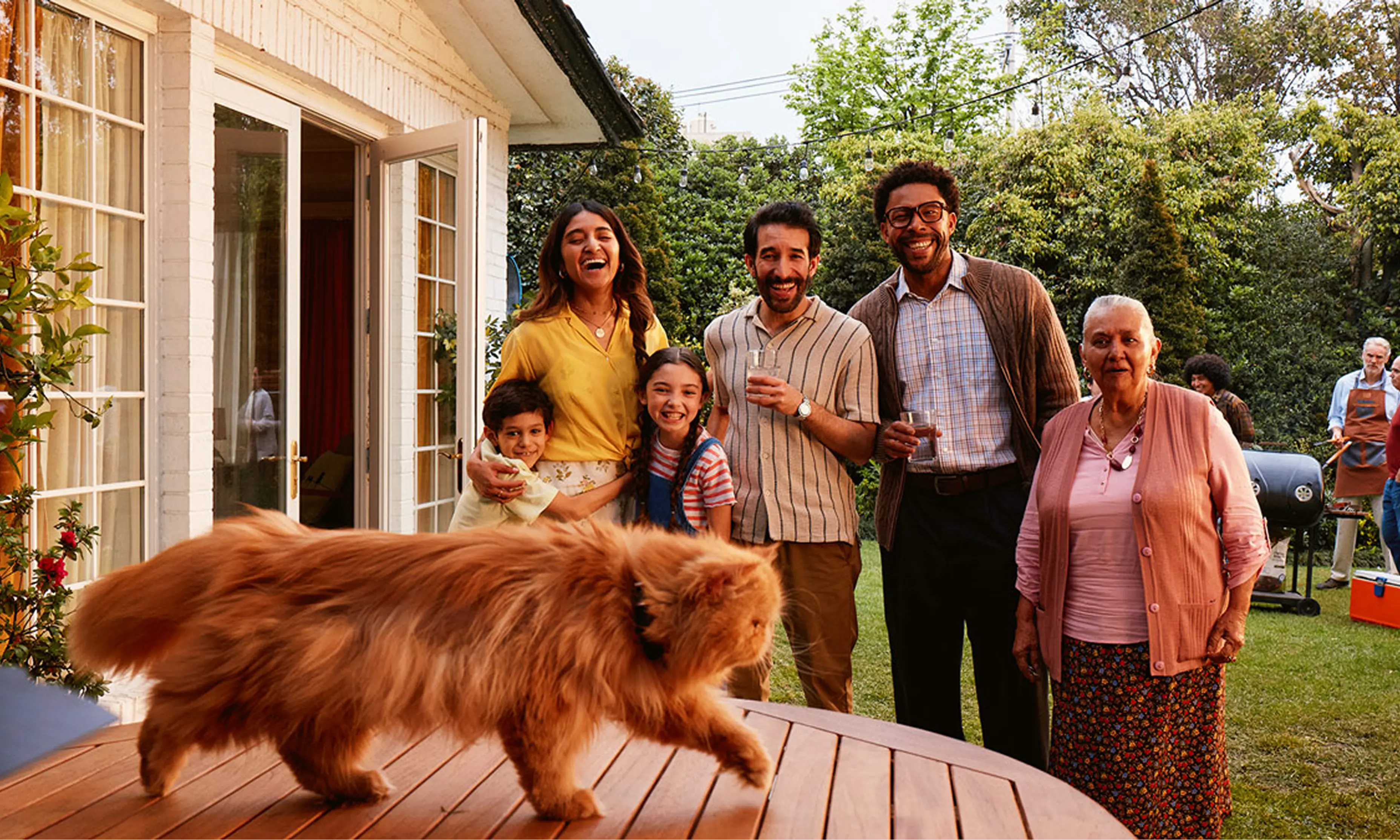 A family outside at a barbecue laughing as their cat walks across the table