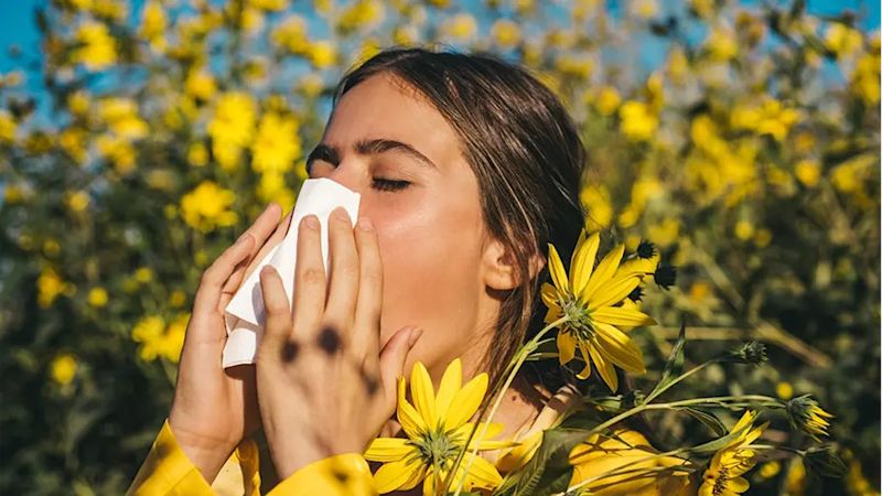  A woman blowing her nose outside surrounded by yellow flowers 