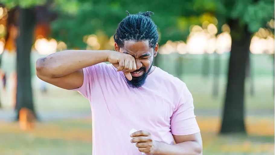 A man in a short sleeve shirt outside, rubbing his eye with one hand, holding a tissue in the other.