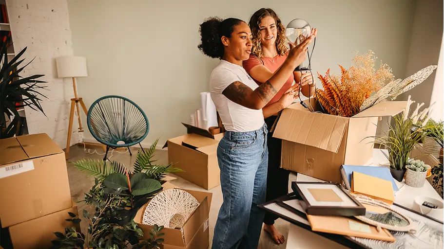 Two women unpack boxes in a room filled with plants, moving boxes, and home decor items.