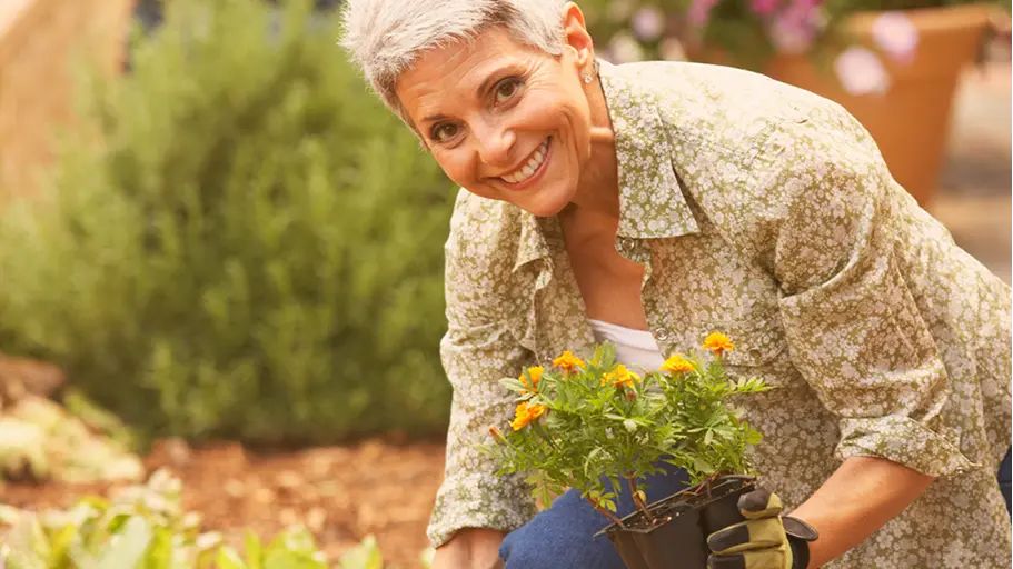 A woman smiles while holding a small potted plant in her hand as she works in her garden.