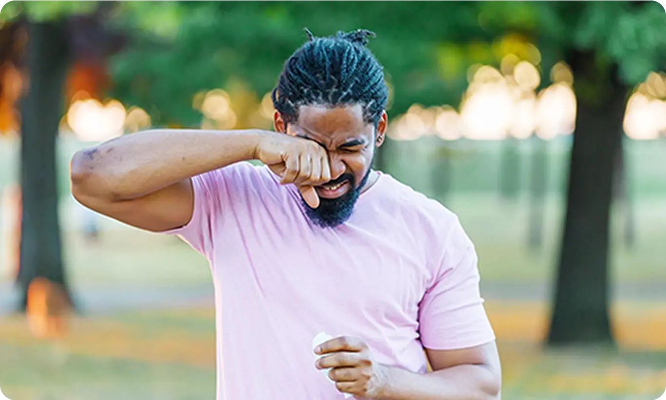 A man outdoors rubbing his eyes and nose, indicating the discomfort of outdoor allergies in a park.