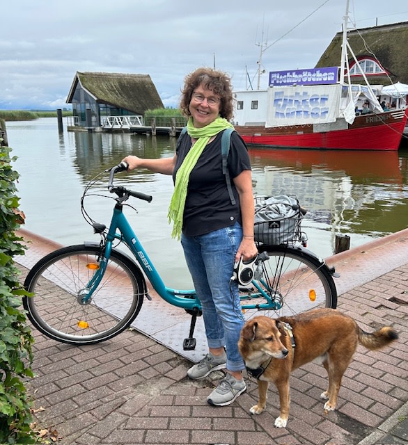 Grundeinkommensgewinnerin Luzi amit Fahrrad und Hund an einem Hafen mit Boot und Reetdächern.