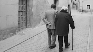 Schwarzweißfoto von zwei alten Menschen, die man von hinten beim Spazierengehen in einer Gasse sieht.