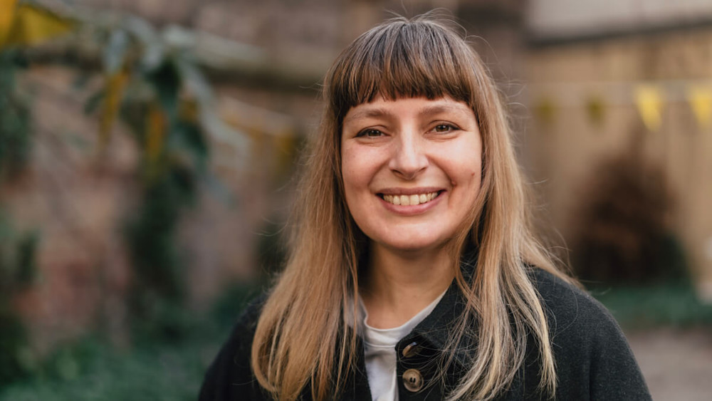 Teilnehmerin Sarah steht lächelnd vor einer Gartenmauer