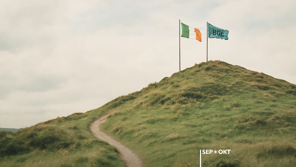 Ein Weg führt einen grün bewachsenen Hügel hinauf auf dessen Spitze eine irische Flagge und eine Grundeinkommensflagge stehen.