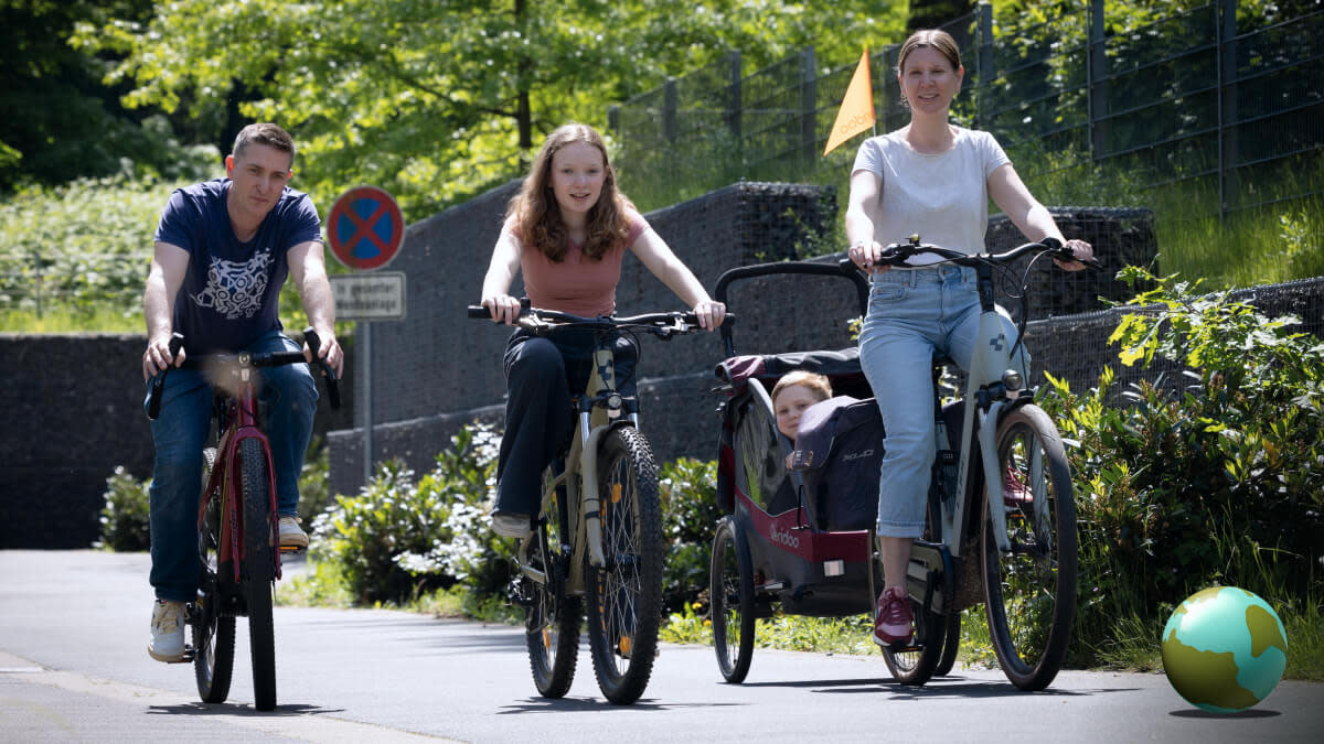 Auf dem Bild sind drei Menschen auf Rädern zu sehen. Sie fahren auf der Straße in Richtung Kamera. Das Fahrrad am rechten Bildrand hat einen Anhänger, da guckt ein Kind raus. 