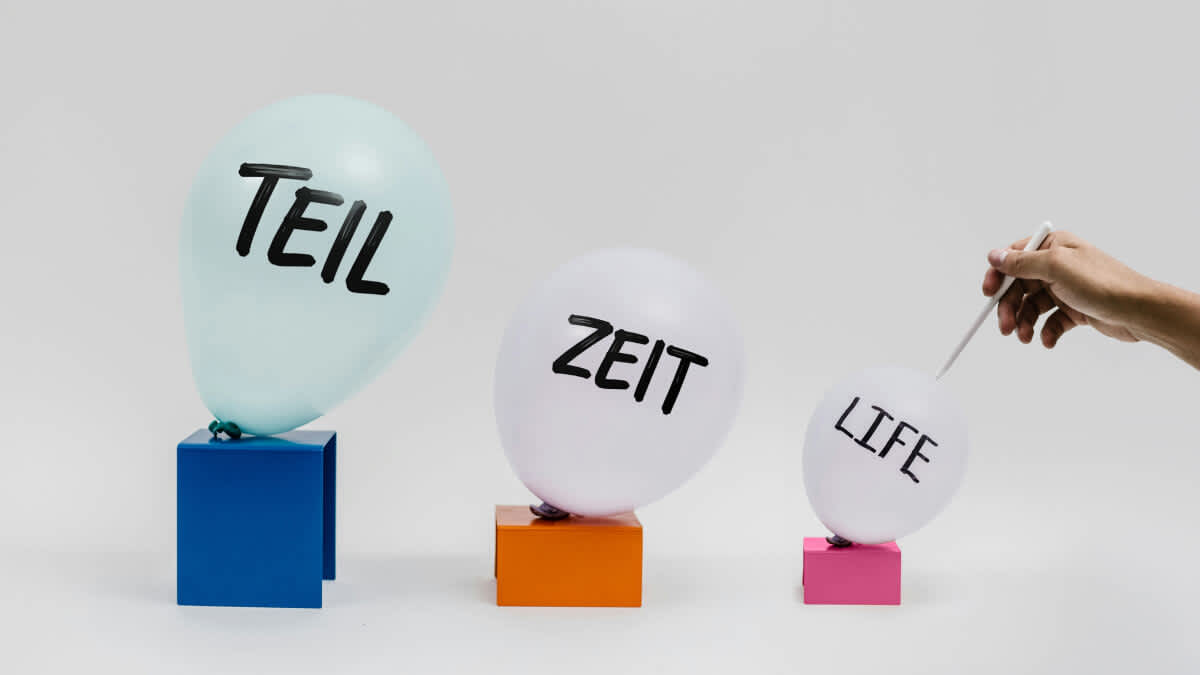 Drei Ballons, auf denen geschrieben steht: "Teil", "Zeit" und "Life". Eine Hand hält eine lange Nadel an den letzten und kleinsten.