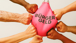 Acht Fäuste boxen auf einen Punchingball, der mit Bürgergeld beschriftet ist.