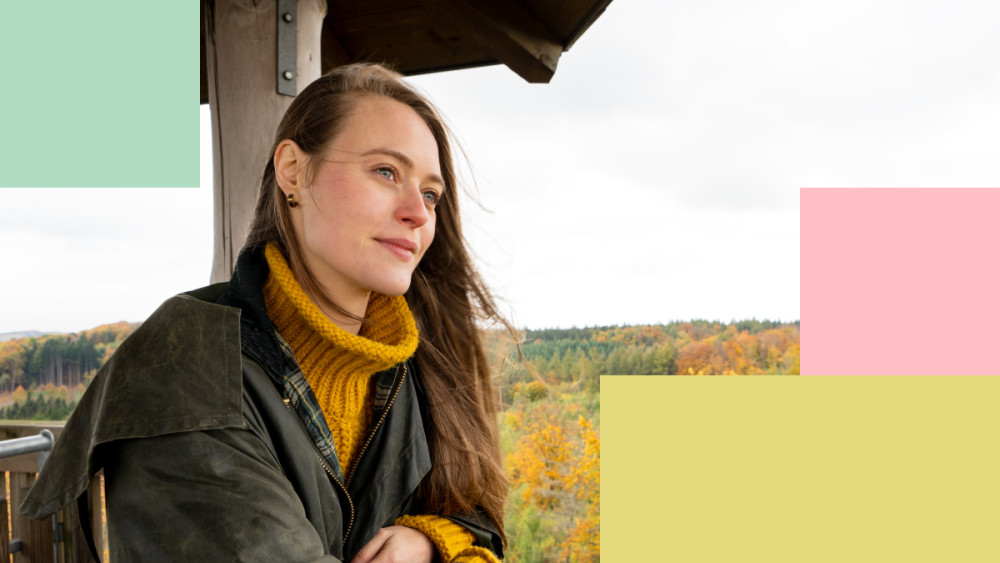 Eine junge Frau steht auf einer Aussichtsplattform und schaut in die Ferne. Im Hintergrund Herbst-Landschaft.