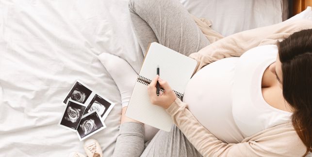 Schwangere Frau sieht Ultraschallbilder an und schreibt Babynamen in ein Notizbuch. 