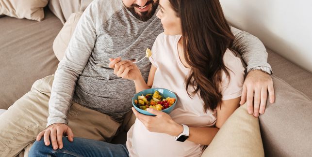 Schwangere Frau sitzt neben ihrem Partner auf dem Sofa und isst einen Obstsalat