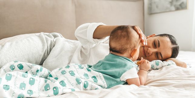 Eine junge Frau liegt mit einem Baby im Schlafsack auf einem Bett und streichelt seinen Kopf.