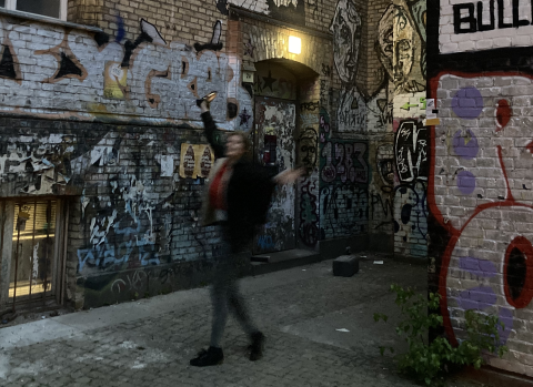 A photo of the entrance to Berlin's rehearsal venue, with a blurry choir member posing joyfully in front of the door. The door is in a brick building covered in grifitti, and the house number 2a is posted next to it.
