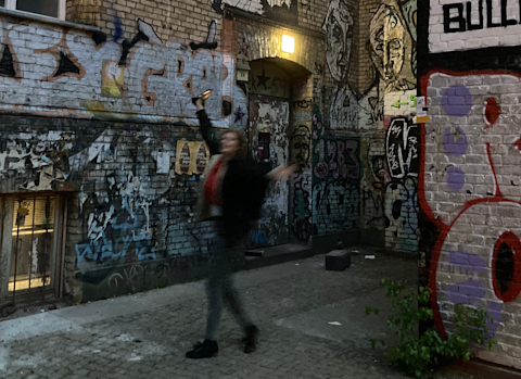 A photo of the entrance to Berlin's rehearsal venue, with a blurry choir member posing joyfully in front of the door. The door is in a brick building covered in grifitti, and the house number 2a is posted next to it.