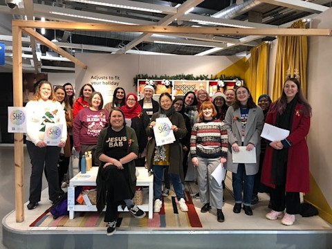 A group of women stand inside the entrance to Ikea. They are in a group, smiling to the camera and holding song lyrics. 