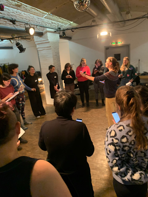 A photo of the Berlin choir rehearsing a song. A semi-circle of choir members is shown, many looking at their phones for lyrics while one choir member has her hands up to lead us. 