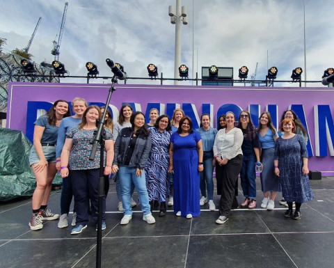 A group of women stand outdoors on a stage, against a purple and silver backdrop. They are all wearing blue clothes and are smiling to the camera. 