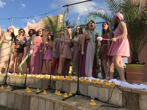 Members of SHE Choir onstage at Secret Garden Party. They are all wearing pink, singing, surrounded by rubber ducks.