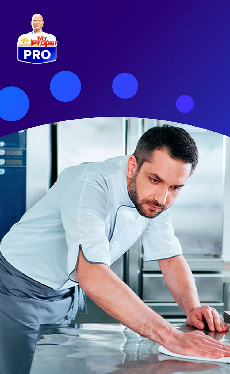 Un chef nettoie un comptoir en acier inoxydable dans une cuisine professionnelle, avec le logo Mr. Propre PRO sur un fond bleu.