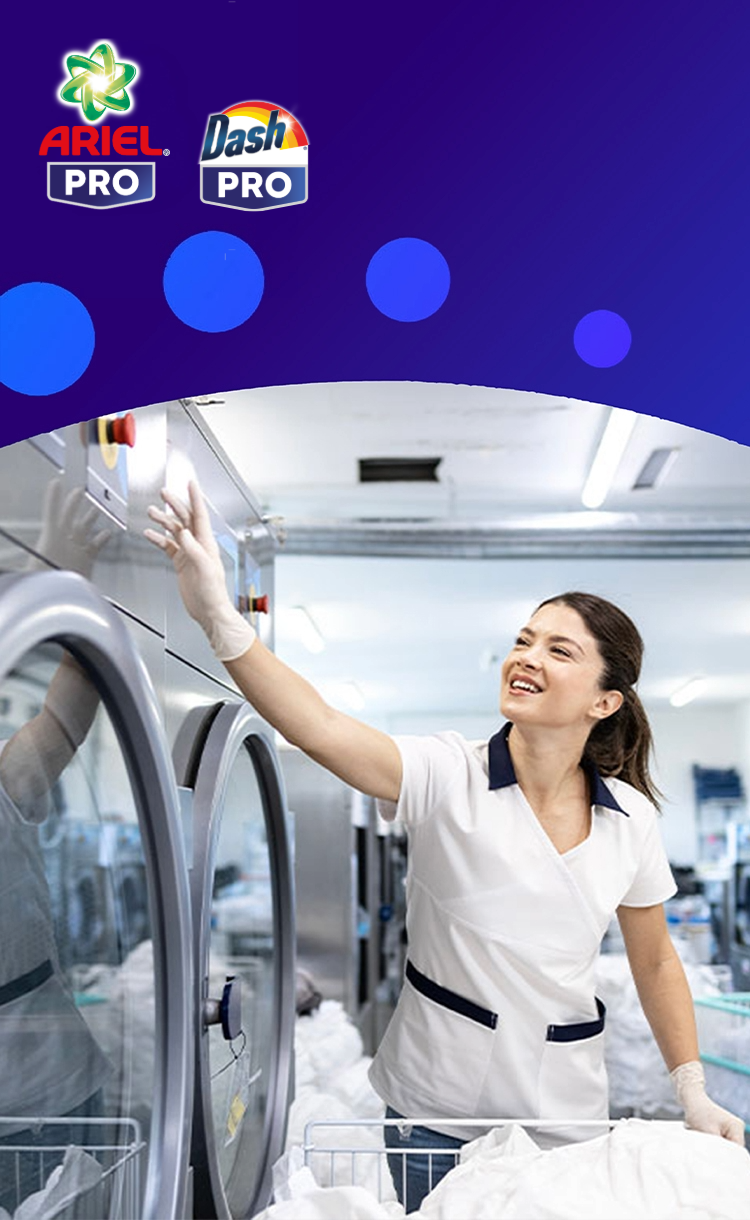 Een lachende vrouw in een wit uniform en handschoenen werkt in een professionele wasserette en reikt naar een rij grote, roestvrijstalen industriële wasmachines. De logo's van Ariel Pro en Dash Pro staan links op een paarse achtergrond.