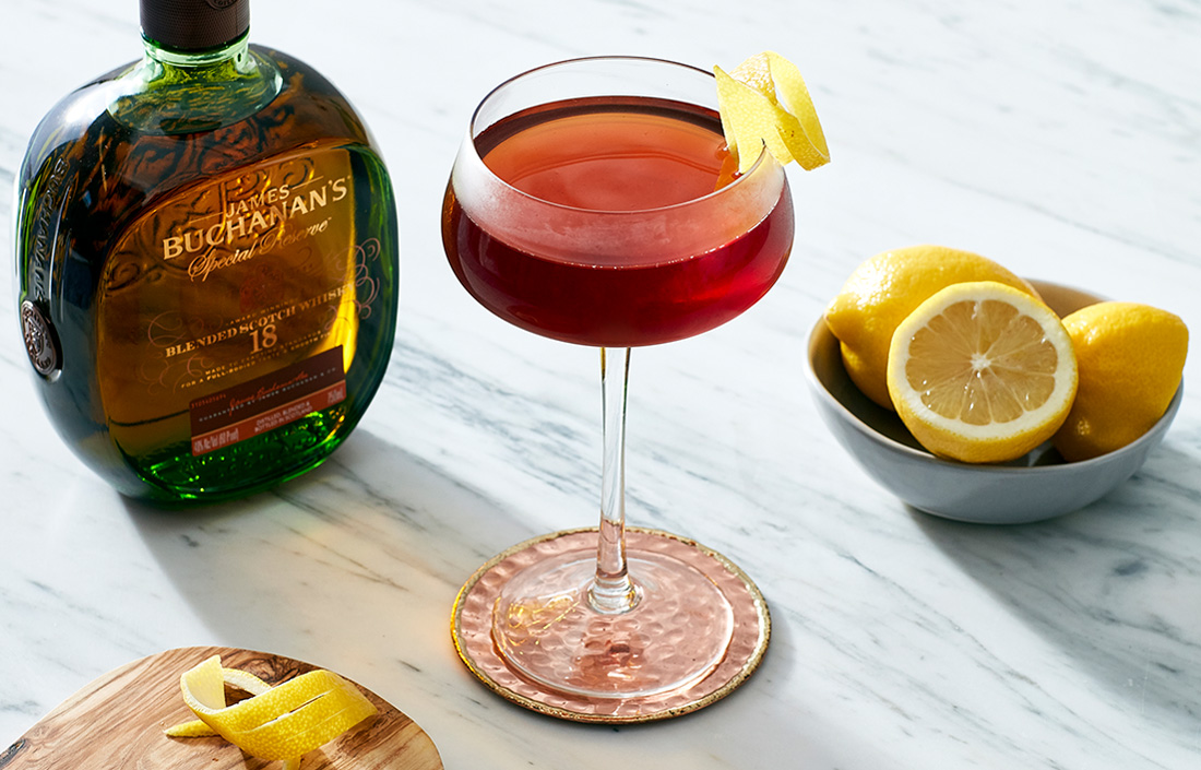 Blender's choice cocktail in a coupe glass surrounded by a Buchanan's 18-year old Special Reserve bottle and a bowl of lemons