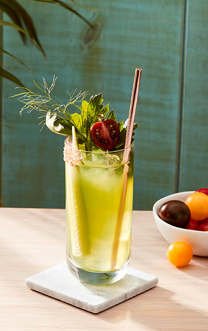 The Buchanan's green bloody marry garnished with a cucumber slice, a celery stalk, and a fresh cherry tomato next to a bowl of fruits